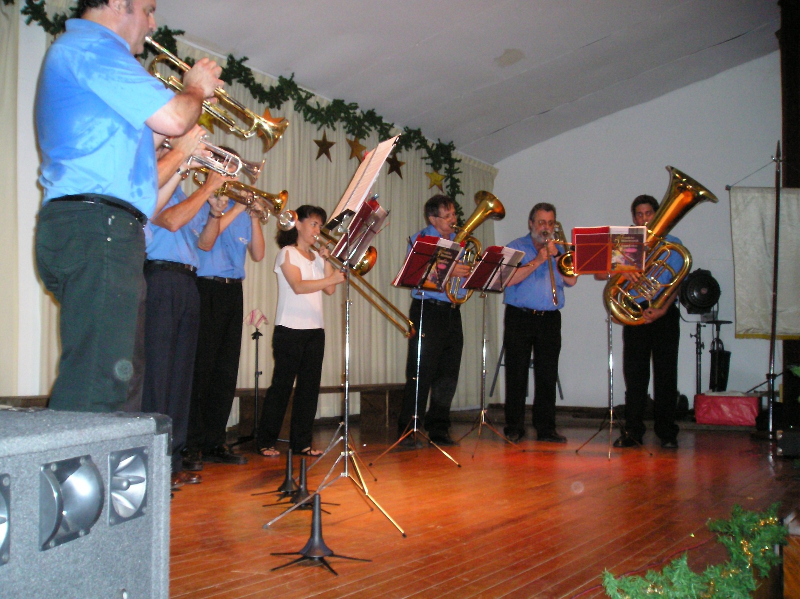 Músicos de camisa celeste tocando de pie sobre un escenario con guirnaldas navideñas