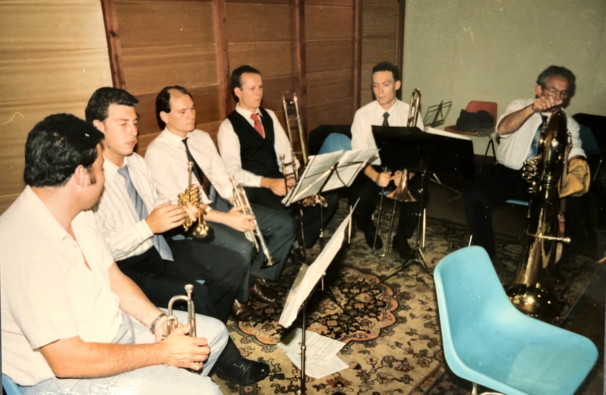 Seis músicos de camisa y corbata sentados en ronda con trompetas, trombón y tuba, frente a atriles, en una sala con paredes de madera y una alfombra