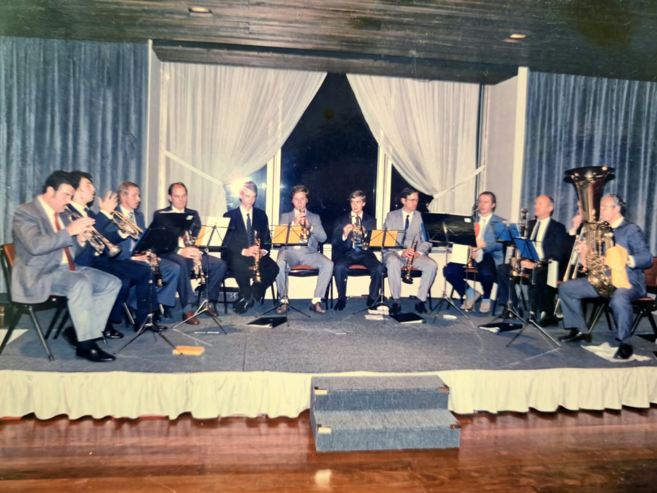 Doce músicos de traje sentados en fila sobre un escenario bajo, con cortinas blancas detrás, tocando trompetas, trombones y una tuba