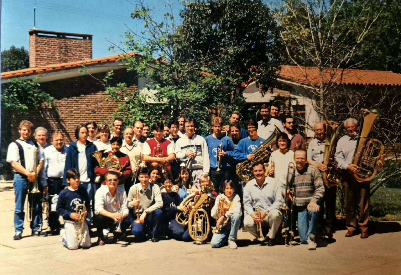 Unos cuarenta músicos con sus instrumentos posando al sol frente a una casa de ladrillo con techo de tejas; adelante, agachados, los más chicos