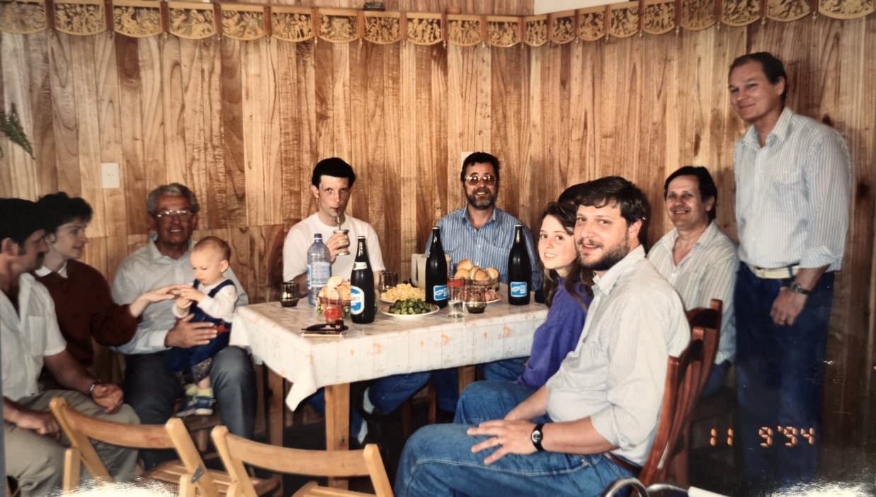 Un grupo alrededor de una mesa con botellas y pan en una sala con paredes de madera; uno toma mate