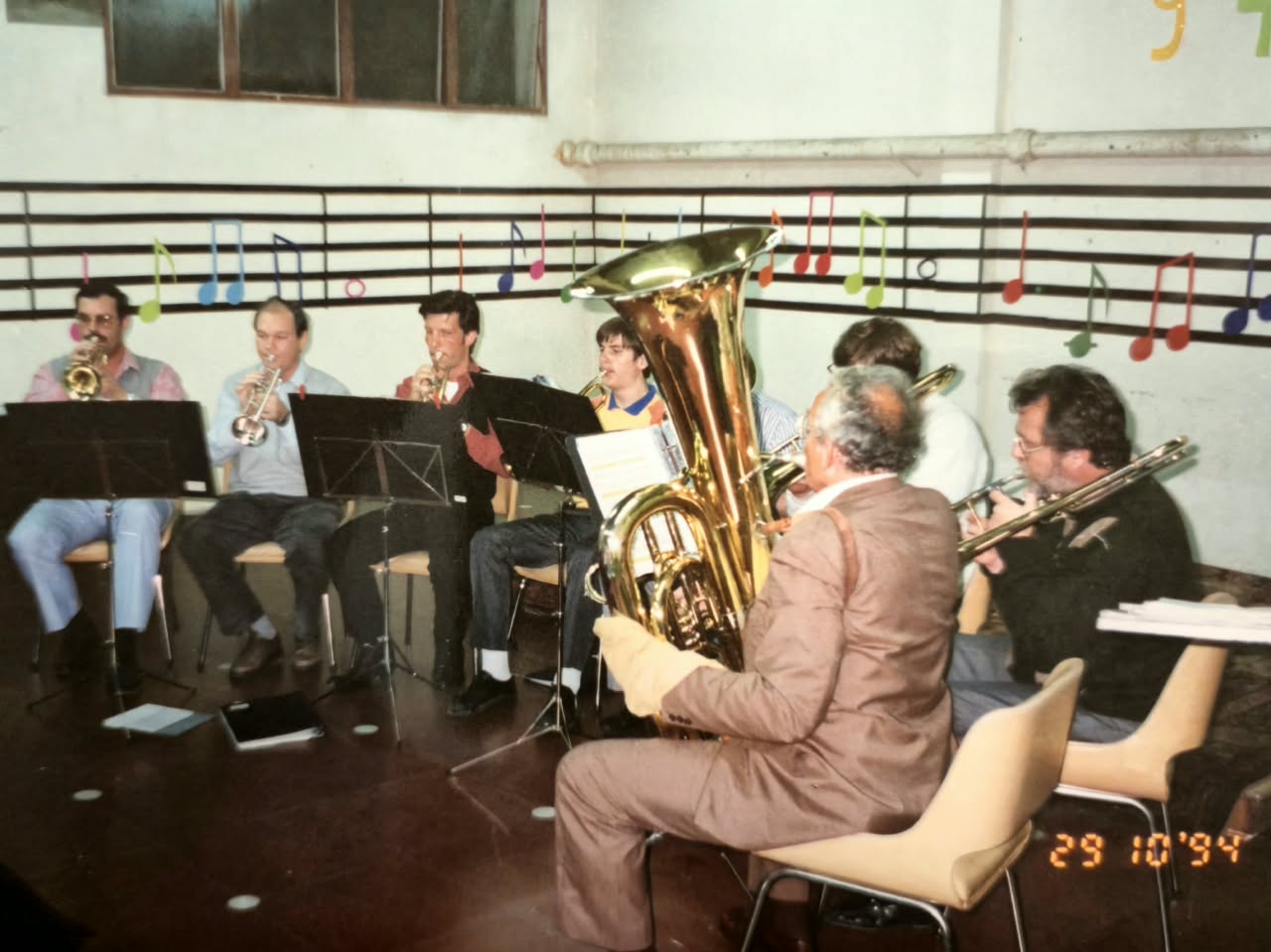 Músicos tocando sentados en un salón con un pentagrama y notas musicales de colores pintados en la pared; en primer plano, un tubista