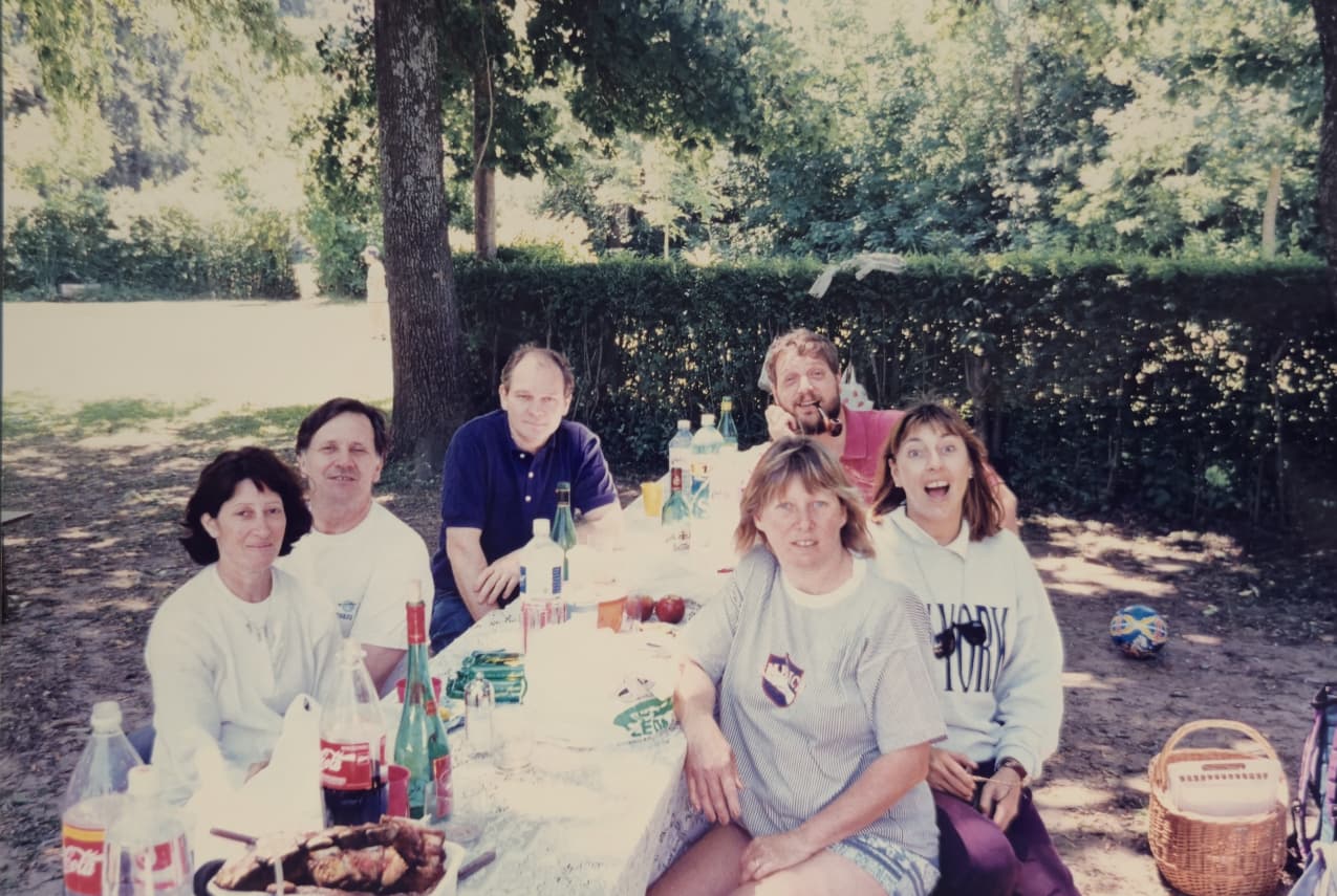Siete personas sonrientes alrededor de una mesa de picnic bajo los árboles
