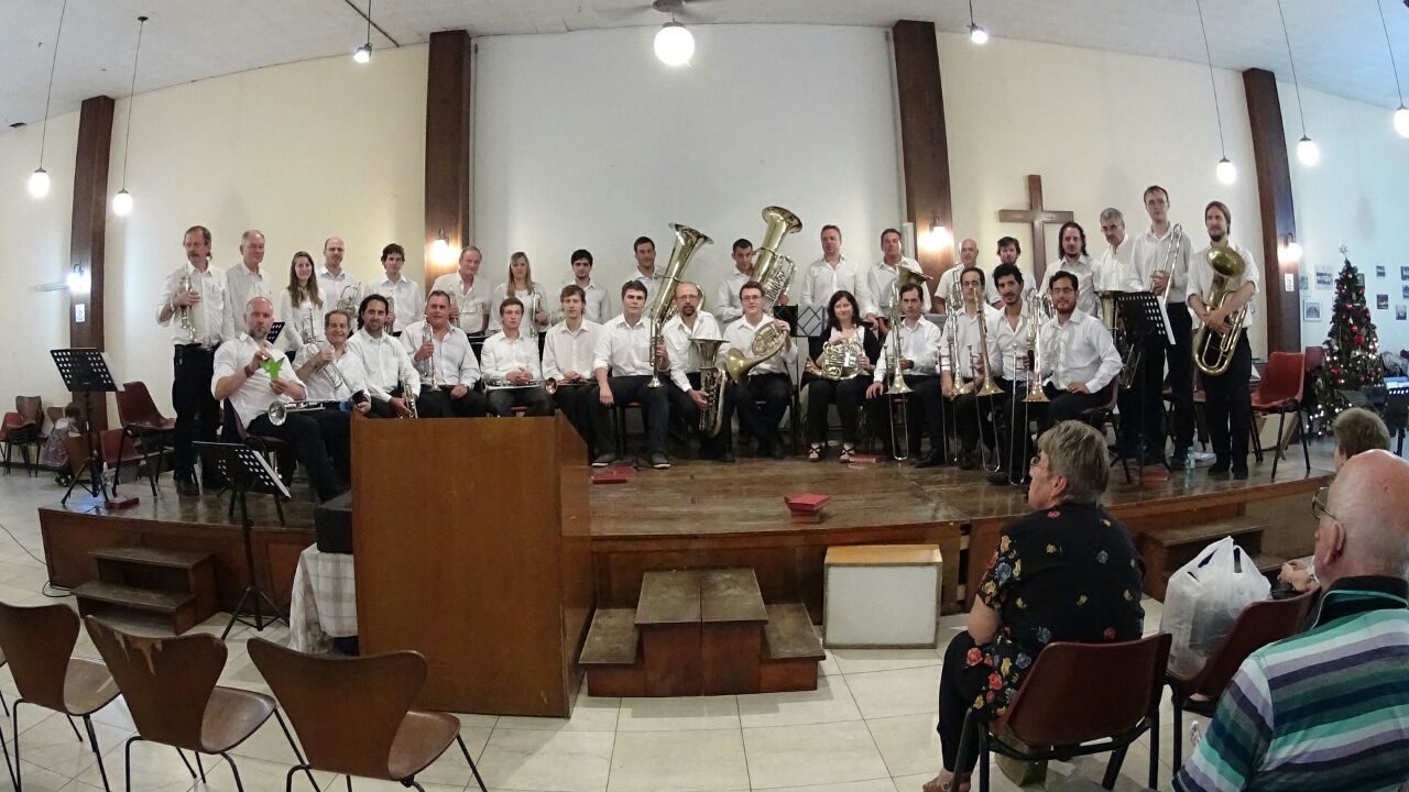 Unos cuarenta músicos de camisa blanca posando con sus instrumentos sobre el escenario de un salón con una cruz de madera y un árbol de Navidad