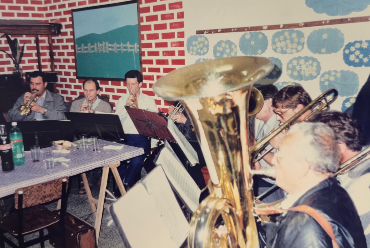 Músicos tocando en un salón de paredes de ladrillo pintado, junto a una mesa con mantel; en primer plano una tuba