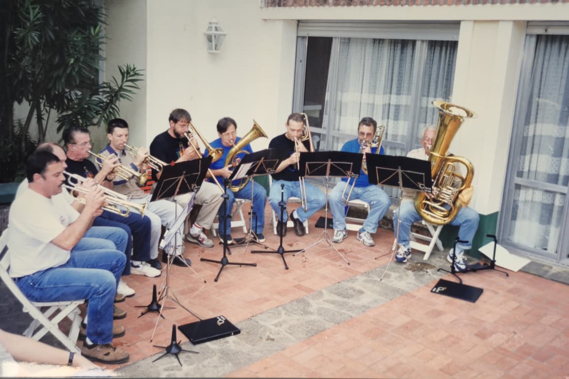 Nueve músicos de ropa informal ensayando sentados en un patio de baldosas frente a una casa