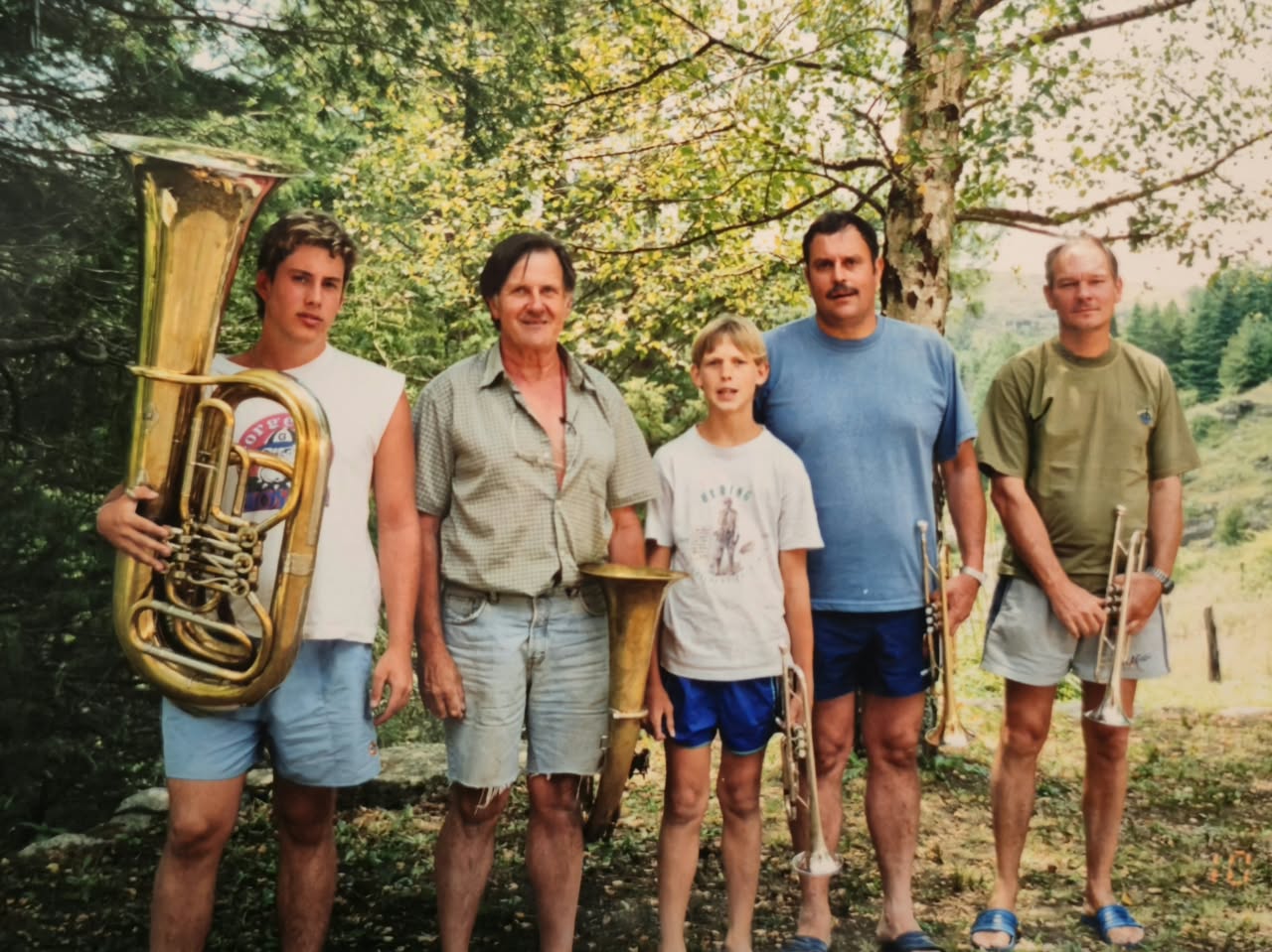 Cinco músicos de ropa de verano posando con tuba, trompetas y trombón entre los árboles de las sierras