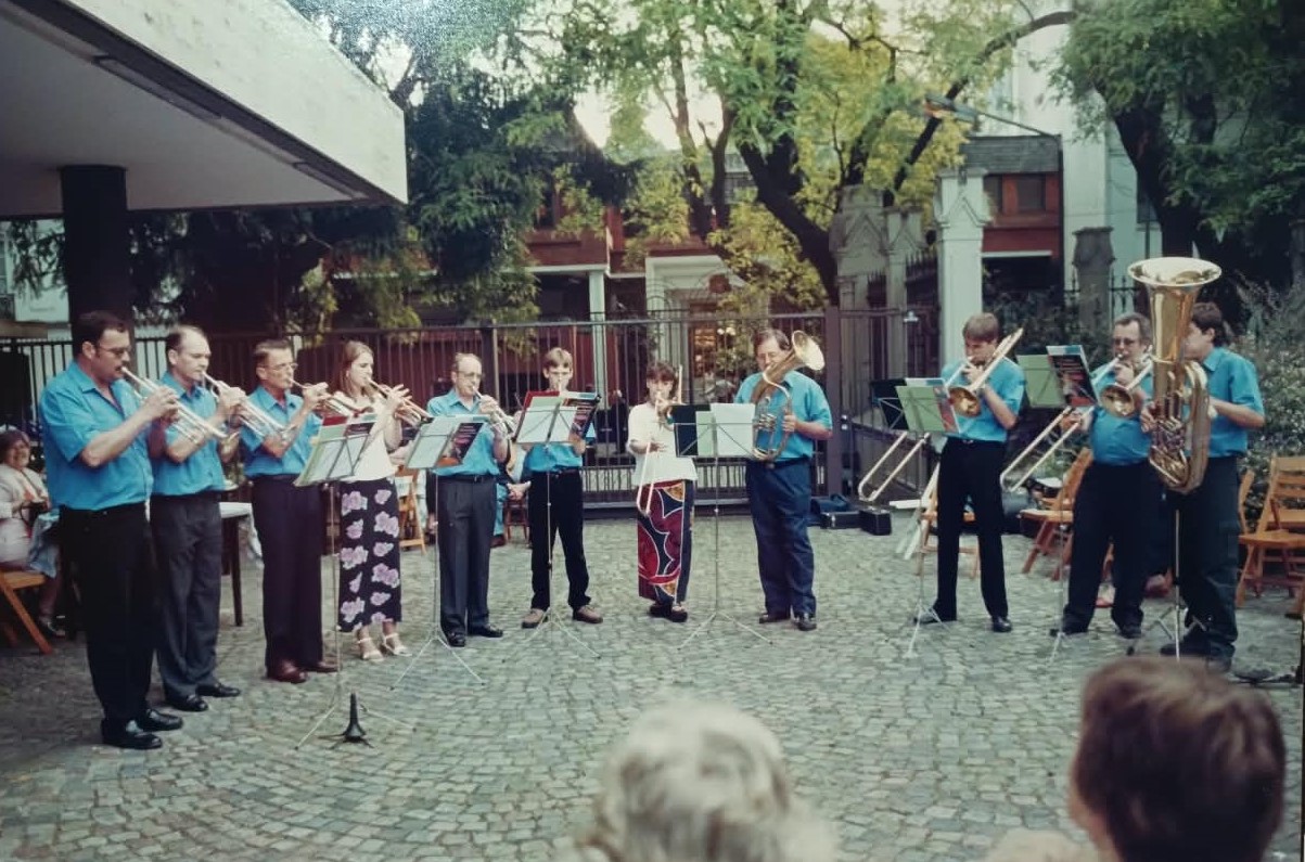 Doce músicos de camisa celeste tocando de pie al aire libre sobre un patio adoquinado