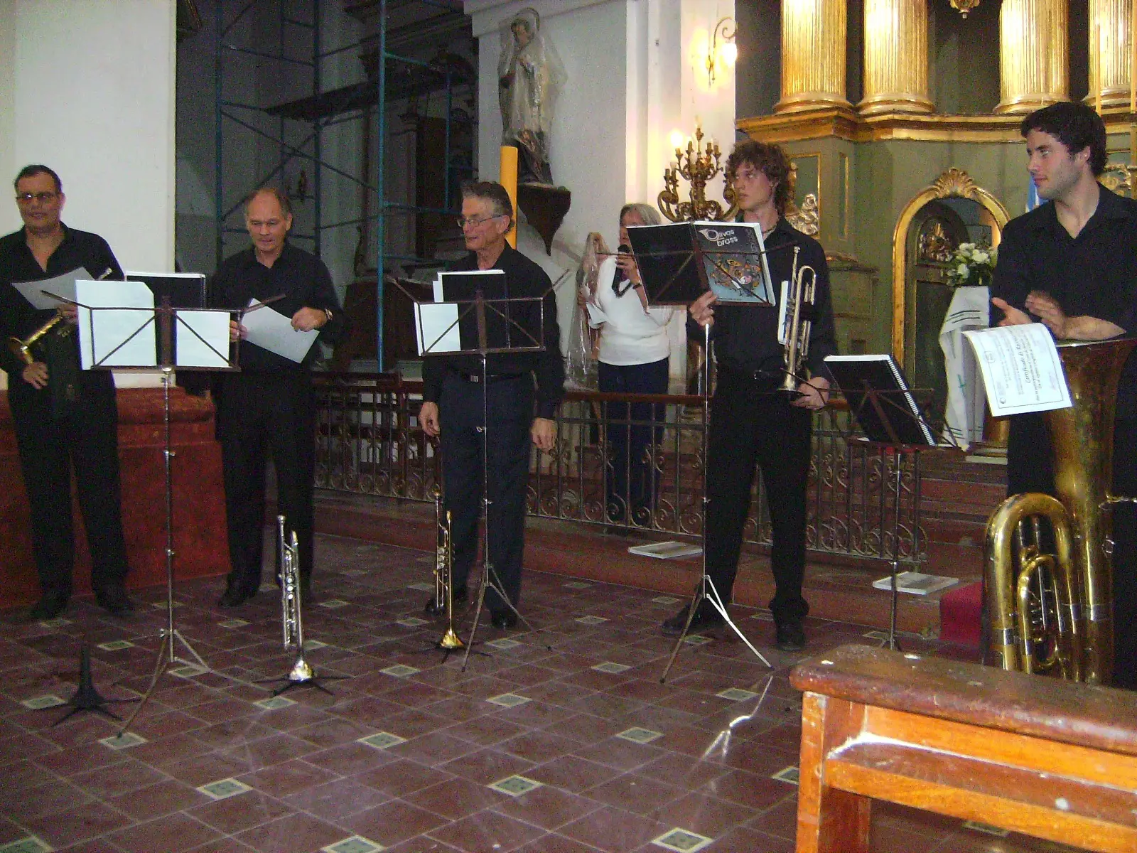 Músicos de negro de pie frente a atriles en una iglesia con altar dorado