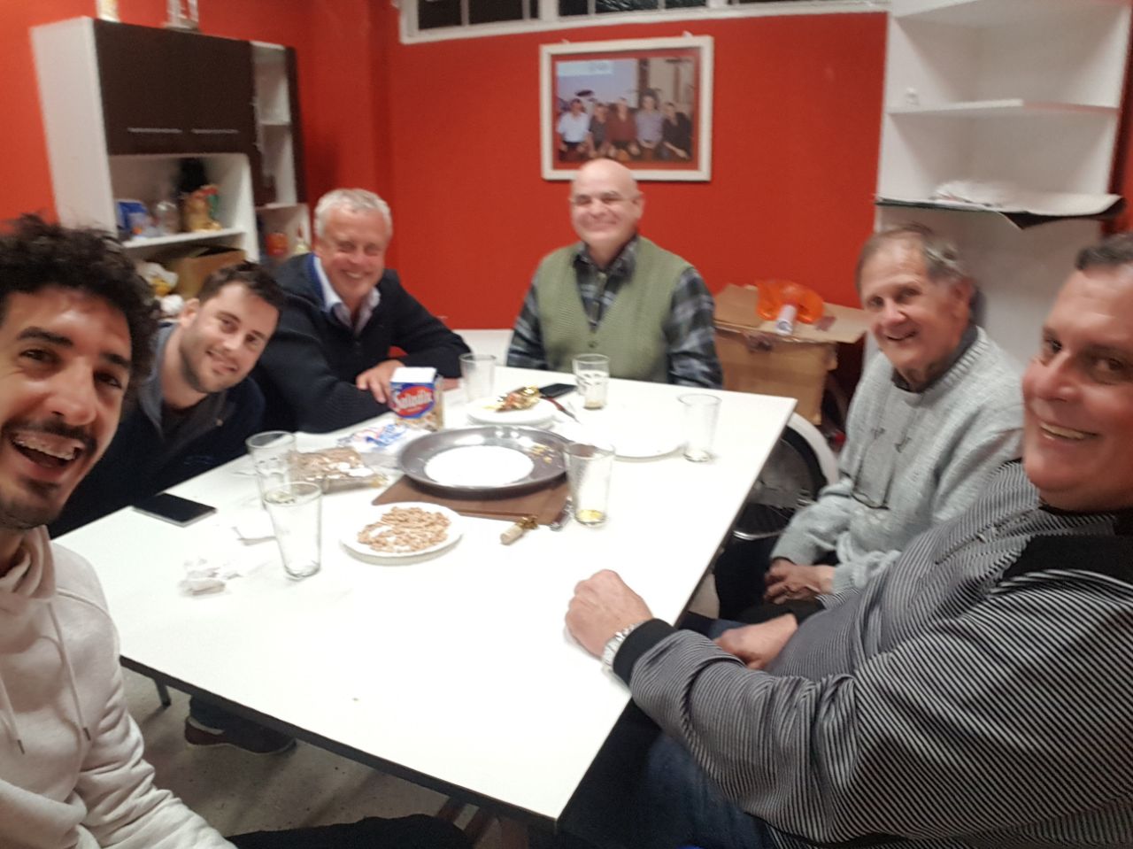 Seis hombres sonrientes alrededor de una mesa en una sala de paredes rojas; el de la izquierda, en primer plano, saca la foto