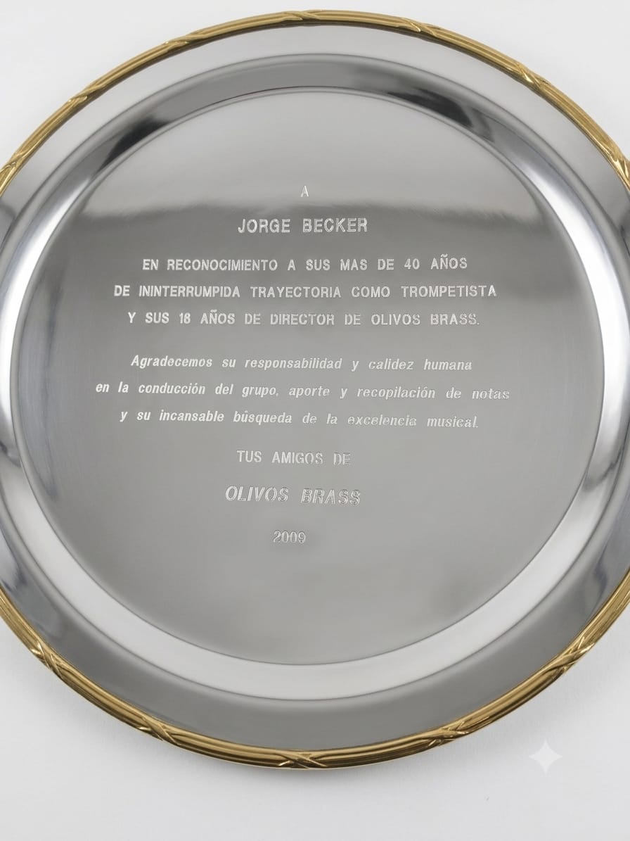 Bandeja circular de metal espejado con borde dorado y texto grabado: «A Jorge Becker, en reconocimiento a sus más de 40 años de ininterrumpida trayectoria como trompetista y sus 18 años de director de Olivos Brass. Agradecemos su responsabilidad y calidez humana en la conducción del grupo, aporte y recopilación de notas y su incansable búsqueda de la excelencia musical. Tus amigos de Olivos Brass, 2009»