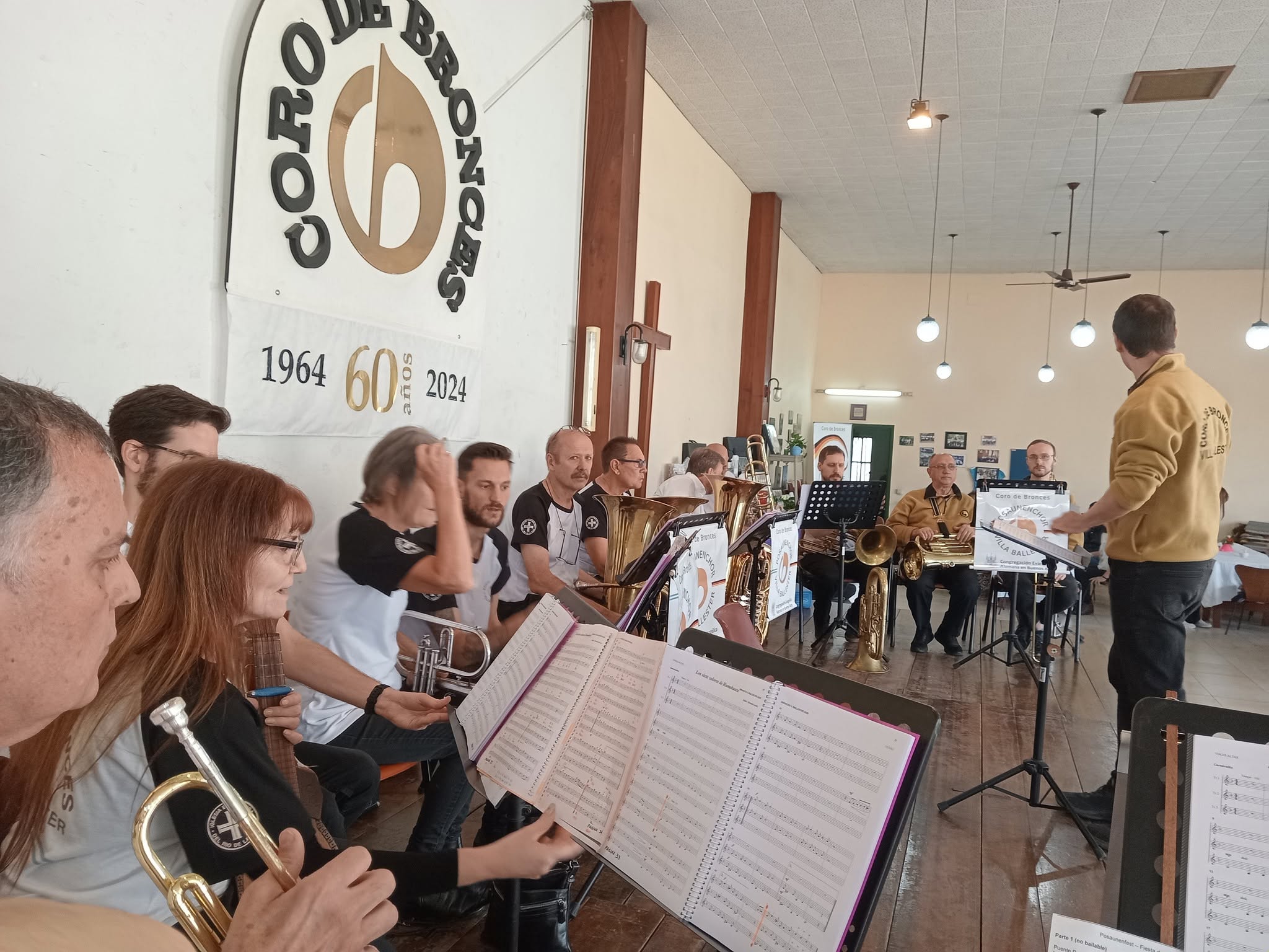 Ensayo del coro en su sala: los músicos sentados con trompetas, tubas y eufonios ante atriles con carpetas de partituras, mientras el director dirige de pie de espaldas; en la pared cuelga el estandarte del coro con la leyenda 1964 · 60 años · 2024