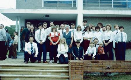 Fotografía en color, de tono virado por el tiempo: unas treinta personas posan en la escalinata y la terraza de un edificio moderno de techo plano con columnas cuadradas; los hombres de camisa blanca y corbata oscura, las mujeres de blusa blanca, cuatro chicos sentados en la primera fila y, al fondo a la izquierda, otras personas mirando entre los árboles; adelante, una tarima de madera y un muro bajo de ladrillo a la vista