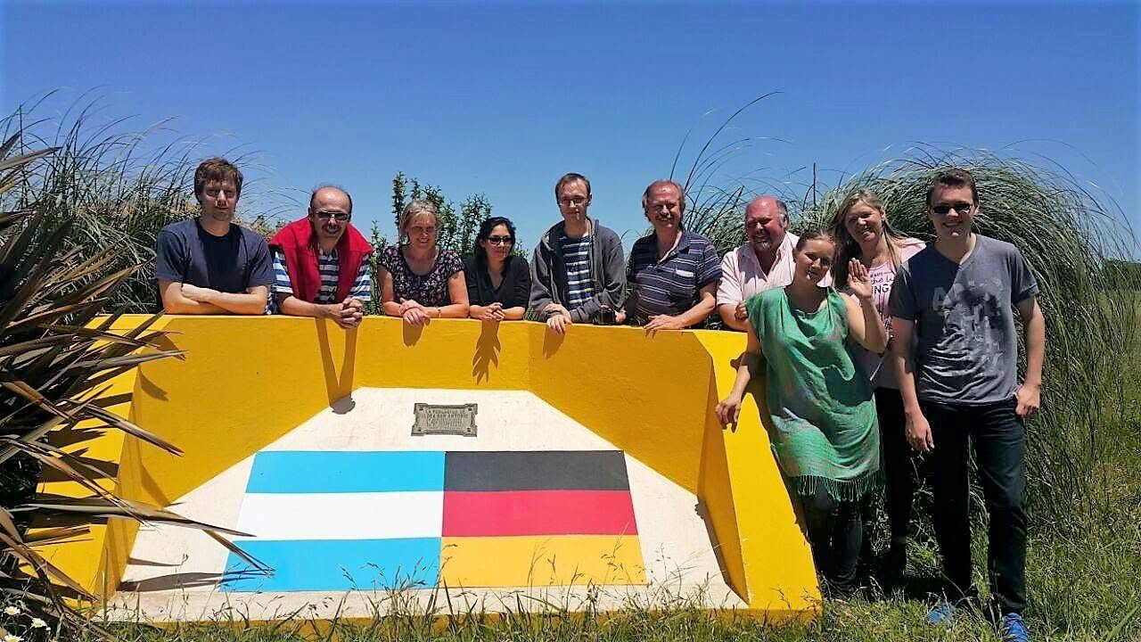 Once personas posan al aire libre apoyadas en un monumento bajo de hormigón pintado de amarillo, con las banderas argentina y alemana pintadas una junto a la otra y una placa de bronce en el centro; alrededor, pastizal alto y cielo despejado