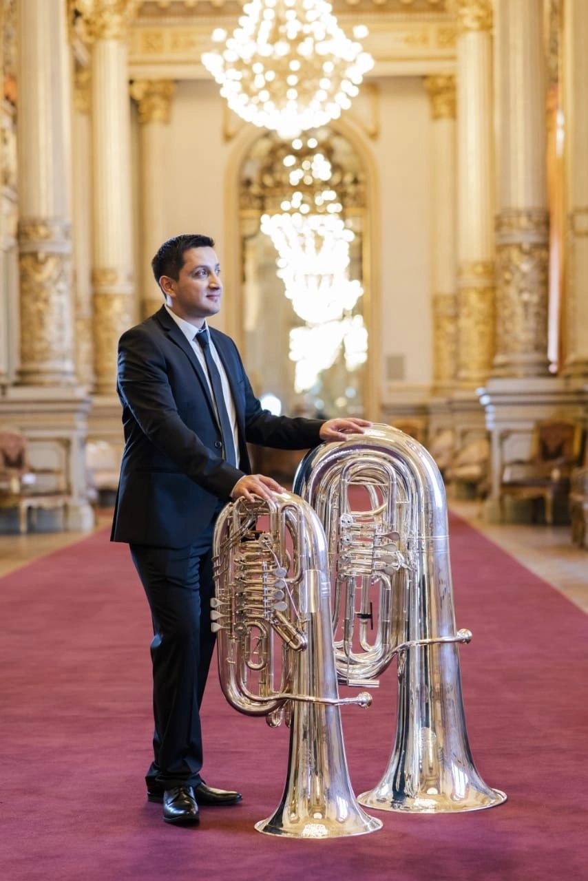 Richard Alonso Díaz de cuerpo entero, traje negro, en un salón dorado con arañas de cristal, la mano apoyada sobre dos tubas plateadas