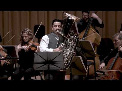 Richard Alonso Díaz tocando la tuba como solista, sentado y rodeado por las cuerdas de la orquesta en el escenario