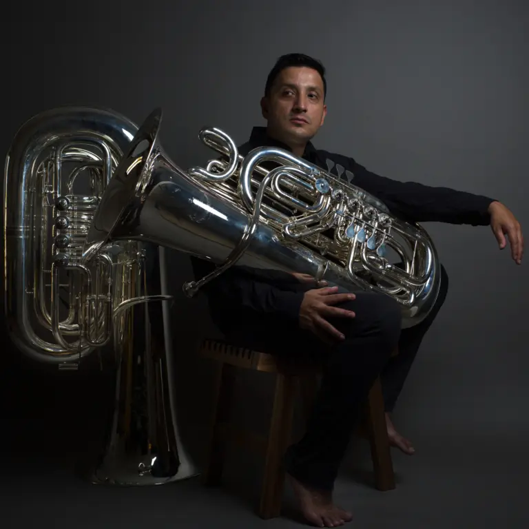 Retrato artístico de Richard Alonso Díaz sentado en un banco de madera, abrazando una gran tuba plateada, sobre fondo gris