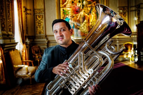 Richard Alonso Díaz con una tuba plateada en un salón dorado con vitral de fondo, sonrisa leve