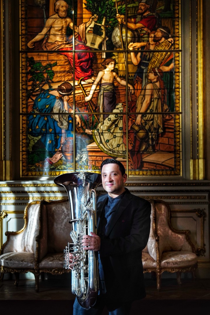 Richard Alonso Díaz de pie, cuerpo entero, con una gran tuba plateada, ante un vitral y sillones antiguos en un salón del Teatro Colón