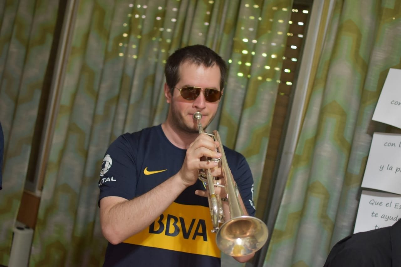 Werner Mengel con camiseta de Boca y lentes de sol espejados toca la trompeta ante una cortina verde