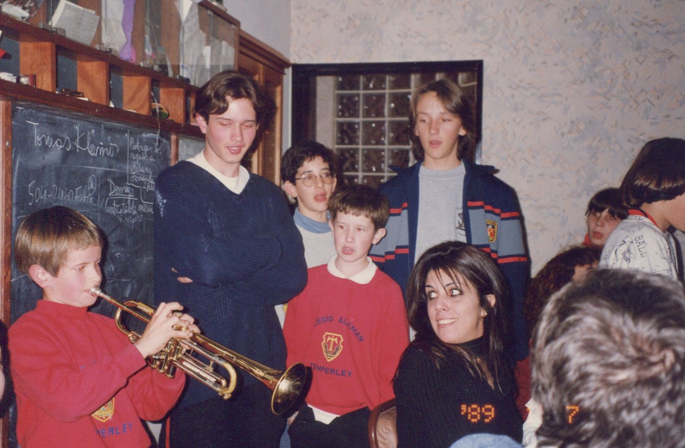 Un chico rubio con buzo rojo toca la trompeta en un cumpleaños en una casa, rodeado de otros chicos, foto de los años noventa