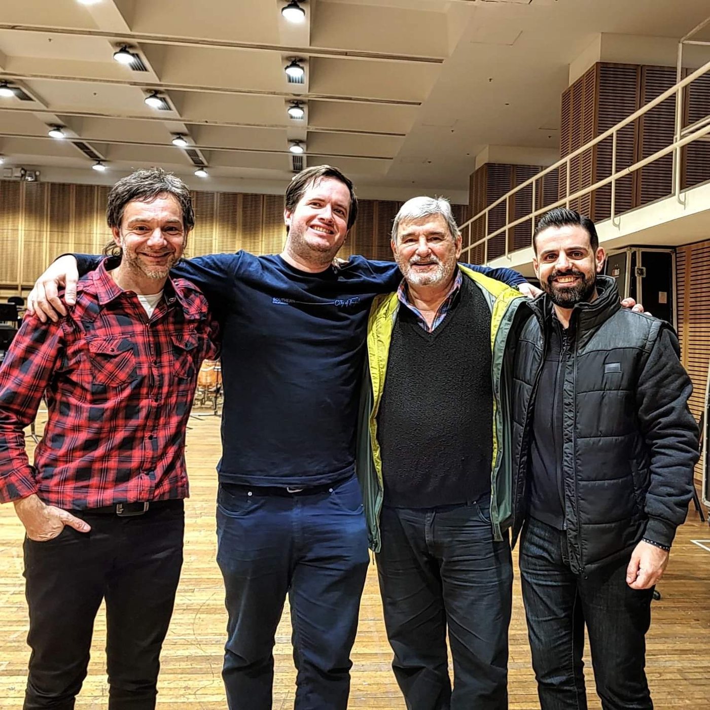 Cuatro músicos abrazados sonríen en una gran sala de ensayo de orquesta con paneles de madera