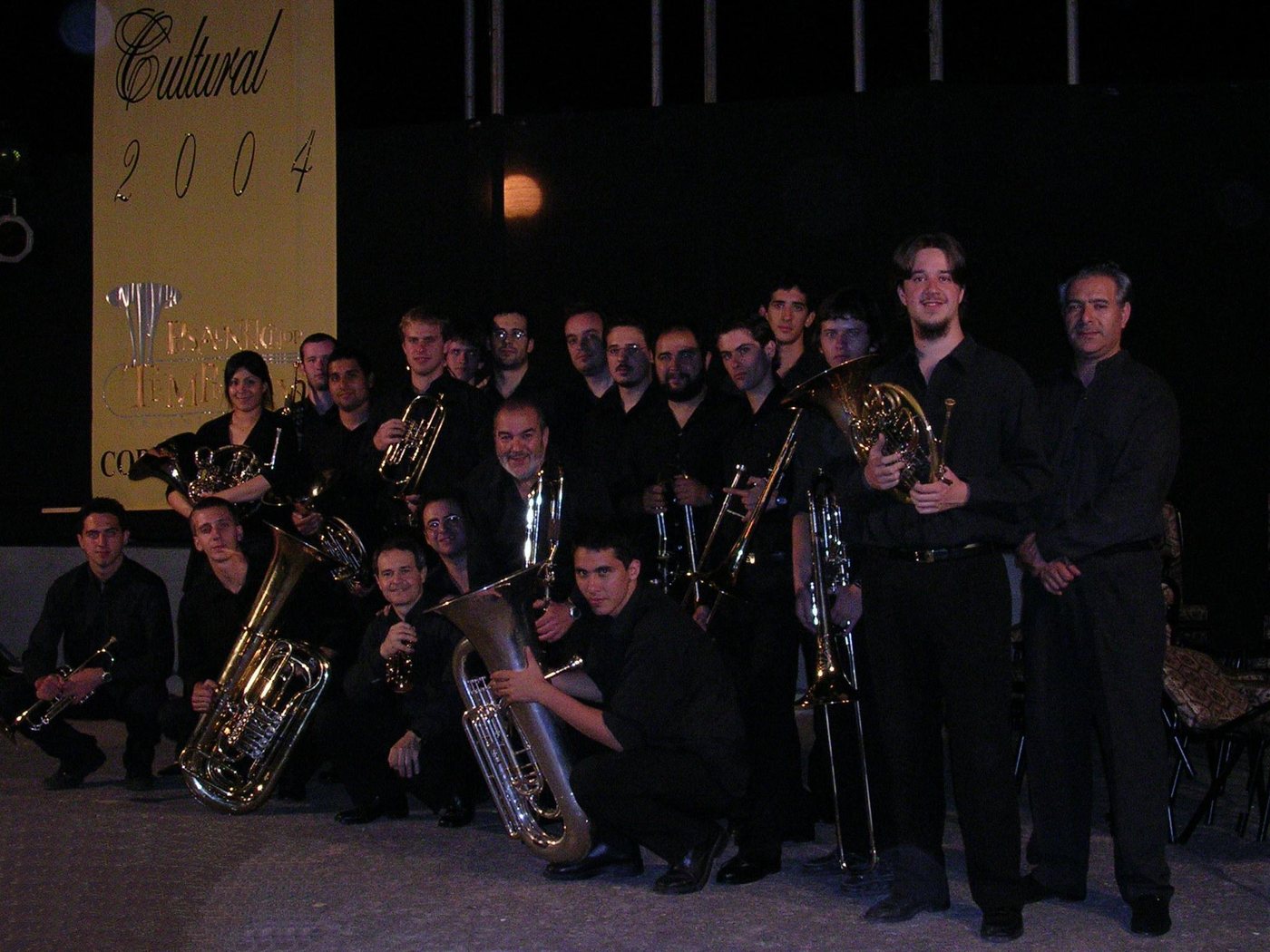 Formación de unos veinte músicos de bronces posando de noche ante el cartel del festival Cultural 2004