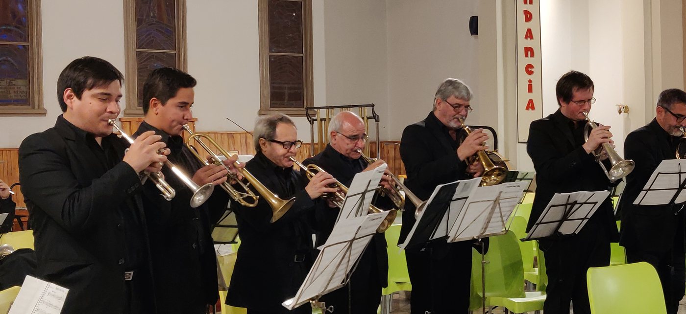 Fila de trompetas y fliscornos tocando de pie en la iglesia Nuestra Señora de la Merced de Villa Ballester