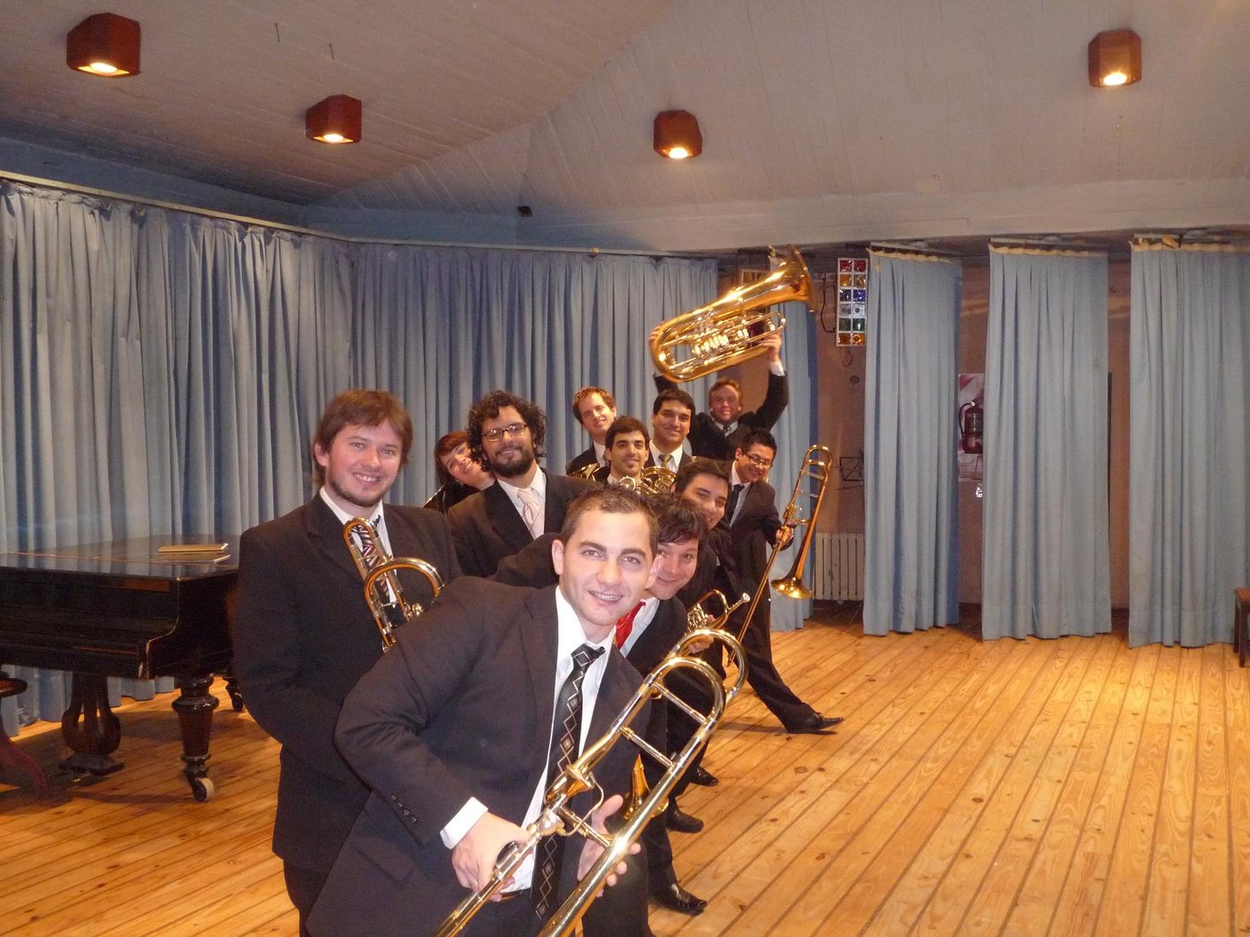 Ensamble de bronces jóvenes de traje posa en fila sonriendo, con un piano de cola al fondo