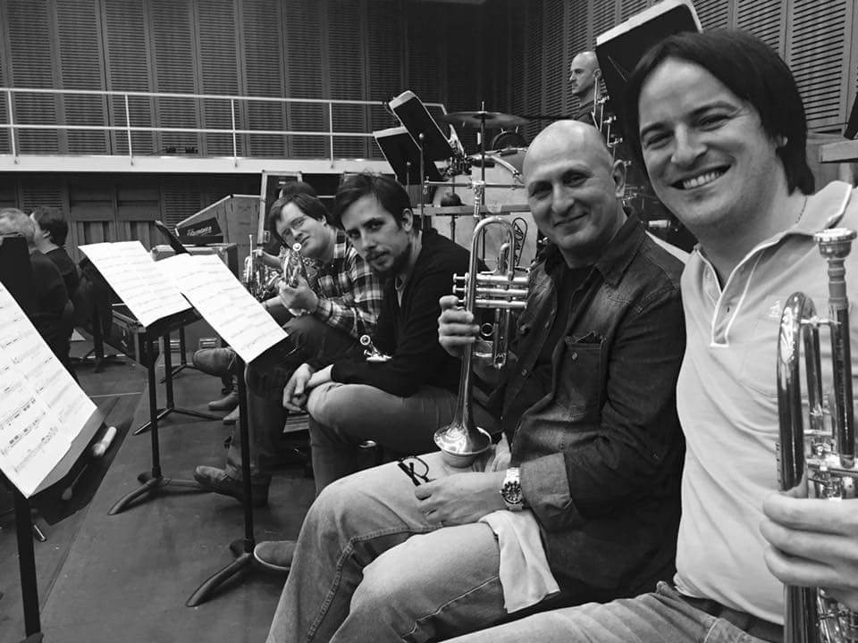 Cuatro trompetistas sonríen a cámara en una sala de ensayo de orquesta, foto en blanco y negro