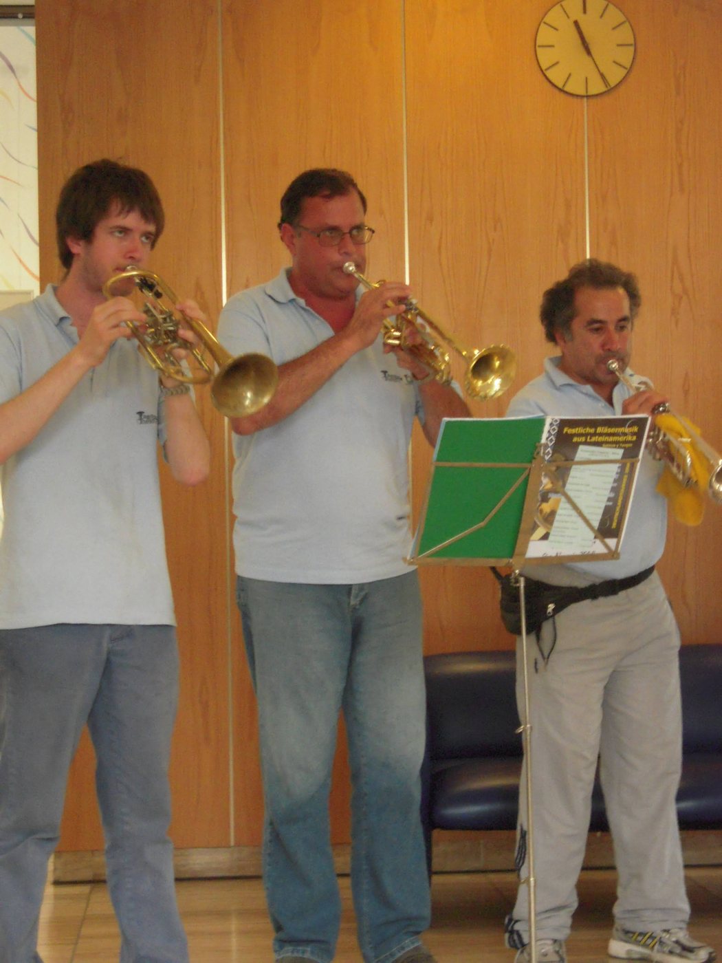 Tres trompetistas de polo celeste tocan ante un atril con el programa Festliche Bläsermusik aus Lateinamerika