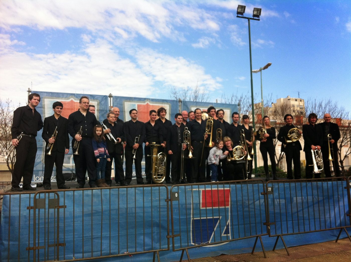 Ensamble de bronces de negro sobre un escenario al aire libre en Lomas de Zamora, con cielo celeste
