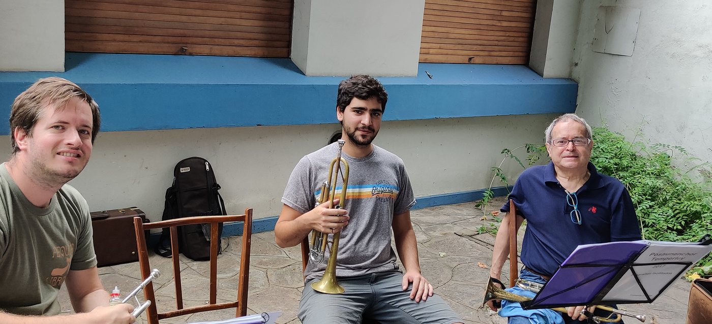 Tres músicos con trompetas ensayan en el patio de la iglesia de Temperley, con una carpeta del Posaunenchor en el atril