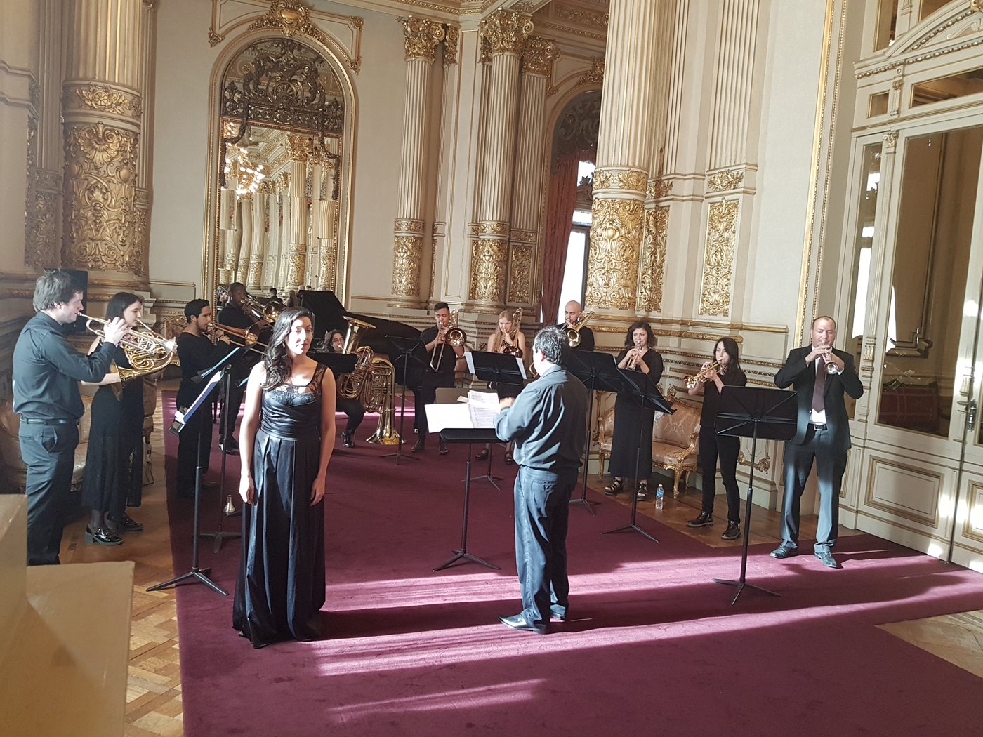 Ensamble de bronces vestido de negro toca con una cantante y director en el Salón Dorado del Teatro Colón