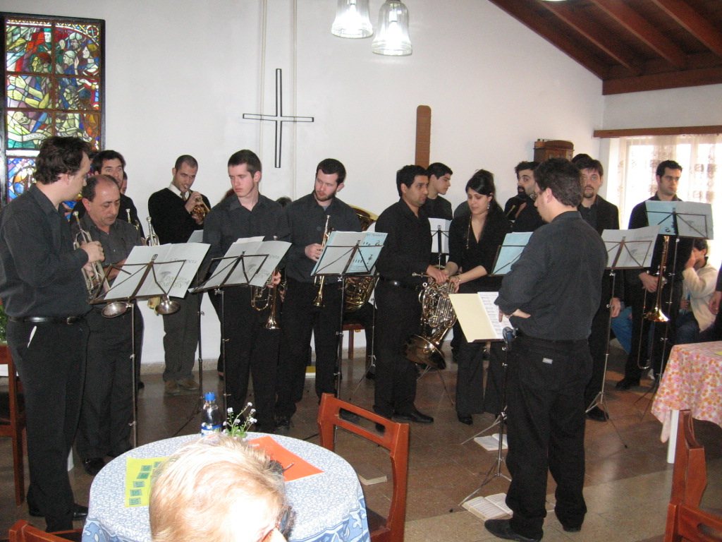 El coro de bronces vestido de negro toca en el templo de Temperley, con el vitral y la cruz al fondo
