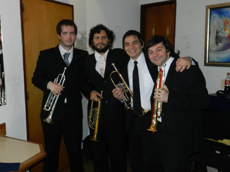 Cuatro trompetistas de traje se abrazan sonrientes en un camarín con sus instrumentos